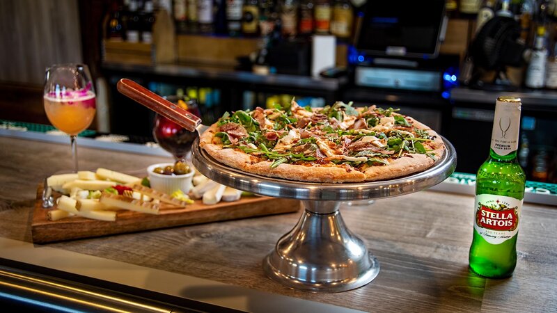 Pizza topped with arugula and prosciutto on bar with a bottle of beer